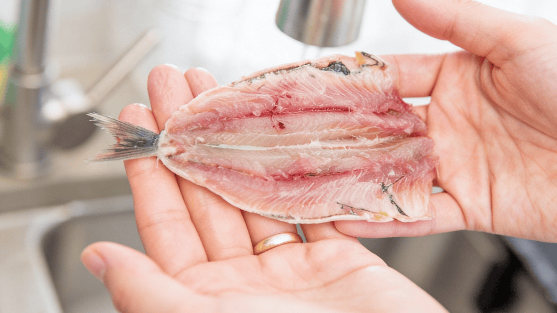 Two hands hold a raw, butterflied fish fillet with the tail still attached over a stainless steel sink.