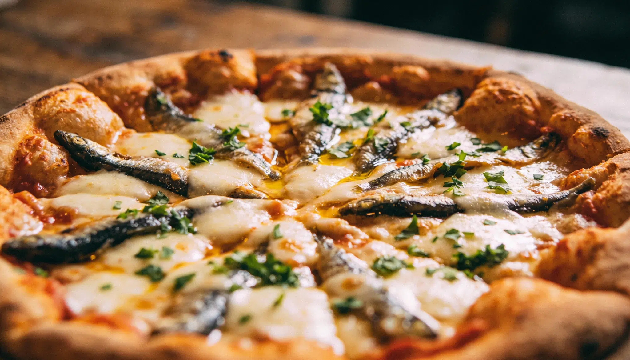A close up of an anchovy pizza with melted cheese, whole anchovies, fresh herbs, and a golden crispy crust on a wooden table.