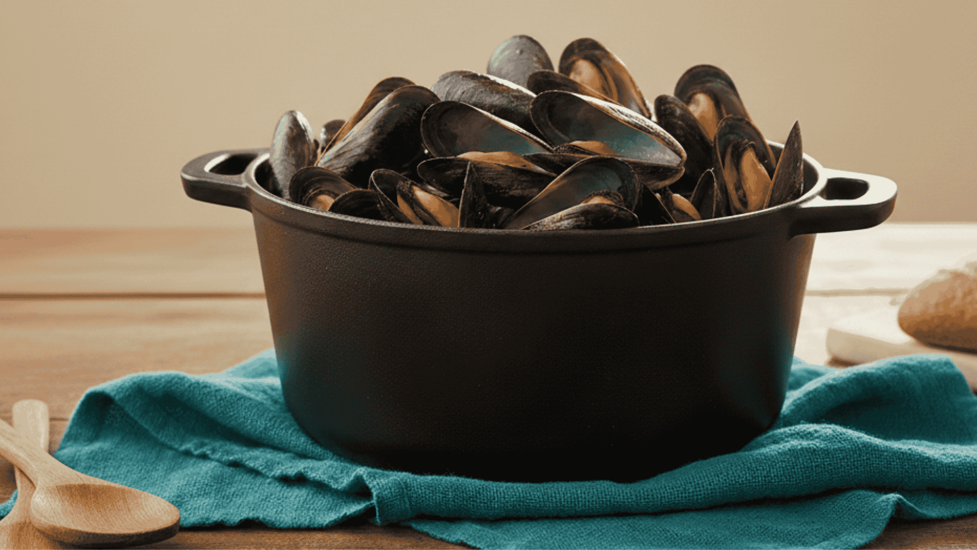 A black cast iron pot overflowing with cooked mussels sits on a teal napkin atop a wooden table