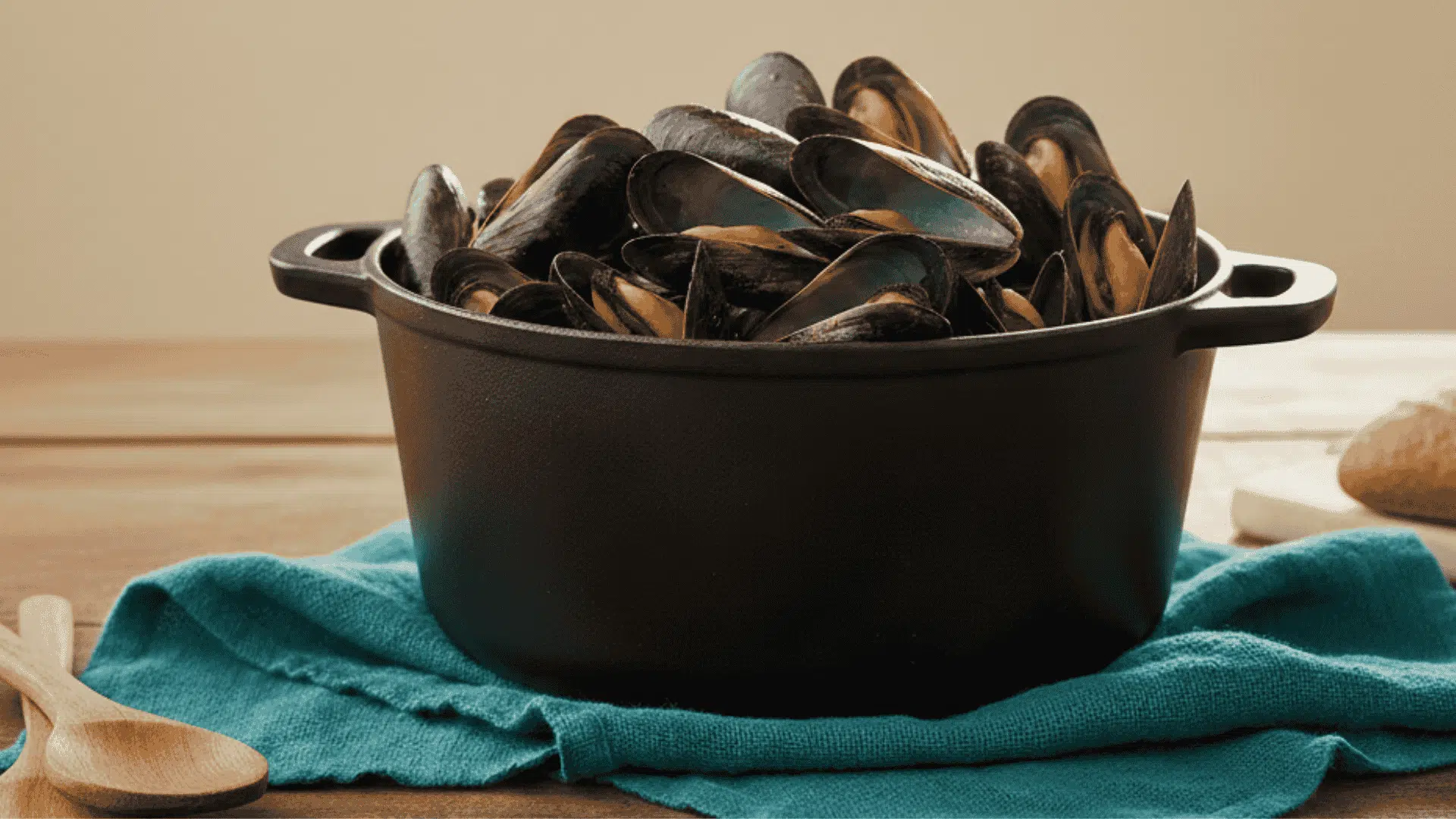 A black cast iron pot overflowing with cooked mussels sits on a teal napkin atop a wooden table