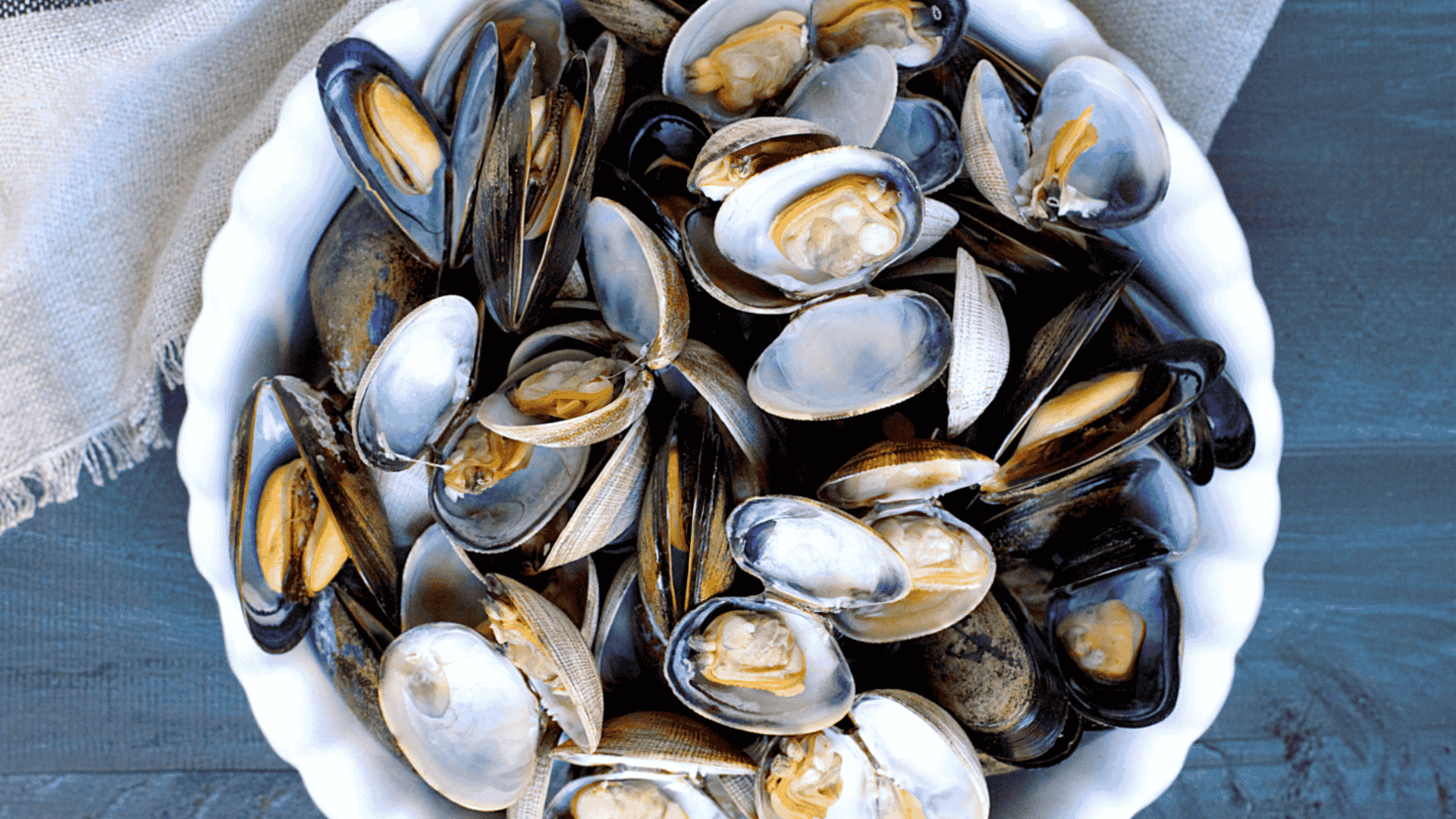 A white bowl filled with steamed and open mussels rests on a textured gray cloth against a dark blue table