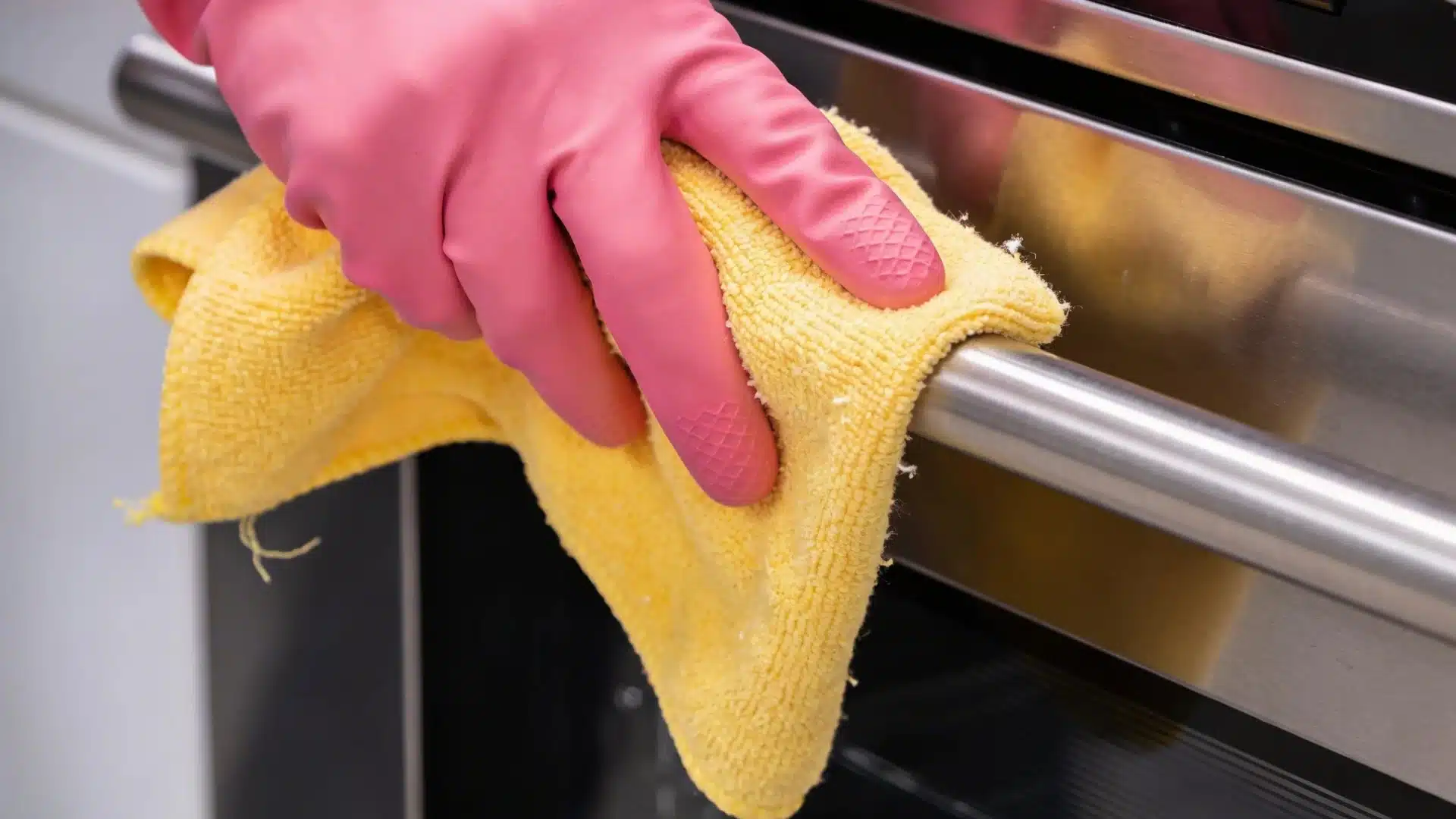  A woman wearing a pink rubber glove wipes the exterior of a stainless steel dishwasher with a yellow microfiber cloth