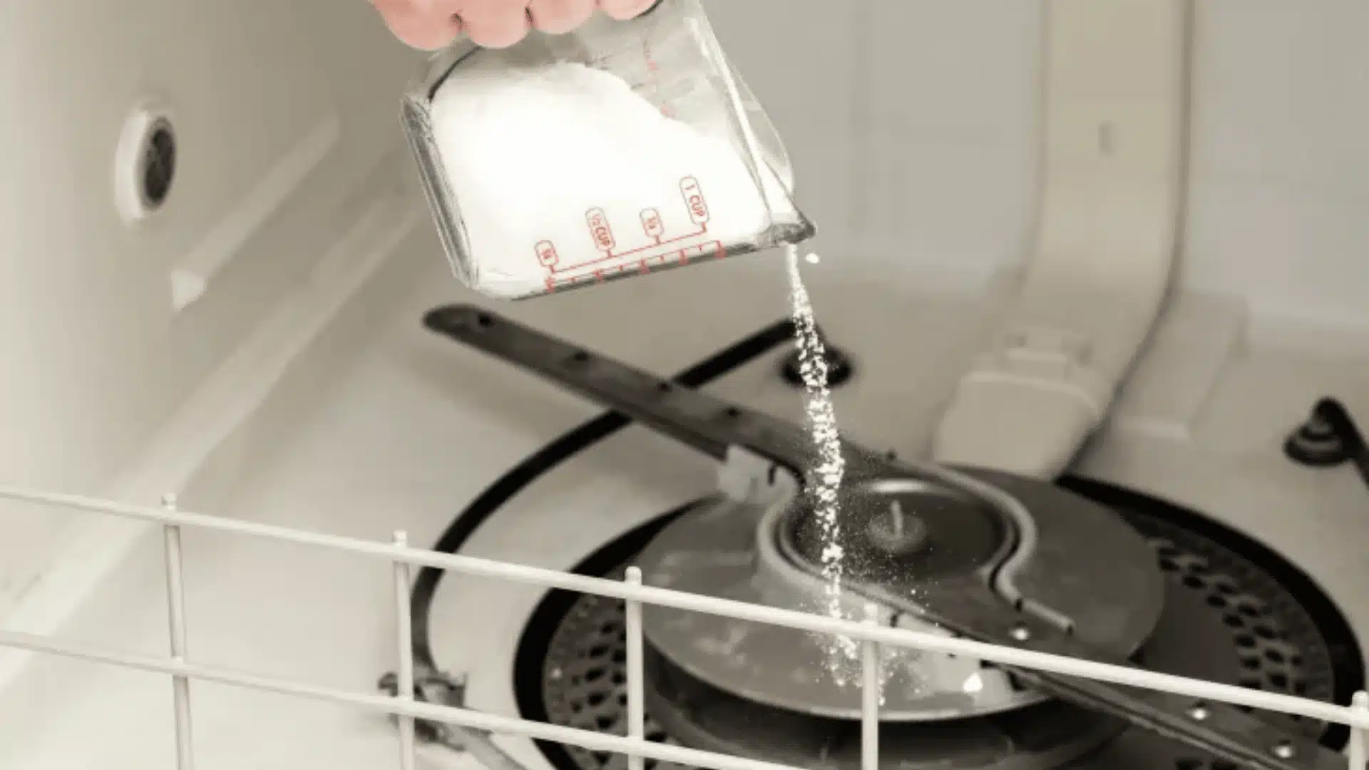 An image of baking soda being poured from a measuring cup into the bottom of the open stainless steel dishwasher