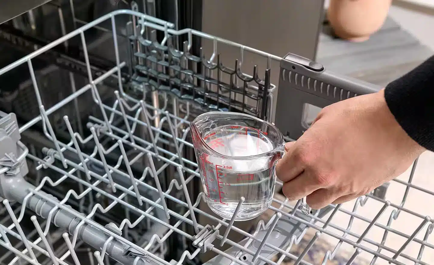 An image showing how to clean dishwasher where a woman is placing a cup of vinegar into the open dishwasher
