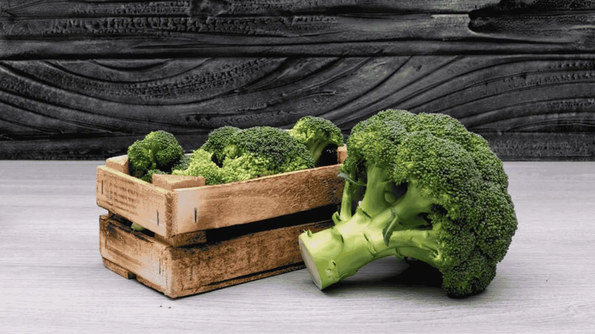Fresh green broccoli florets in and next to a small wooden crate on dark textured wood background