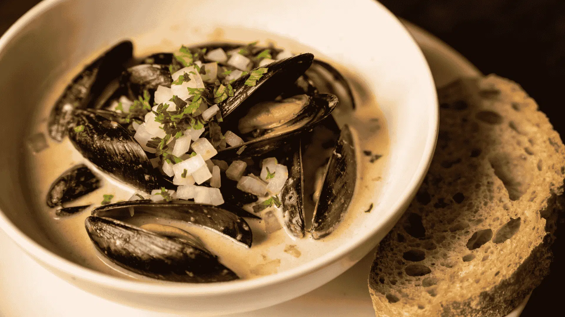 Mussels in a creamy broth, topped with diced white onion and parsley, served in a white bowl next to a slice of crusty bread