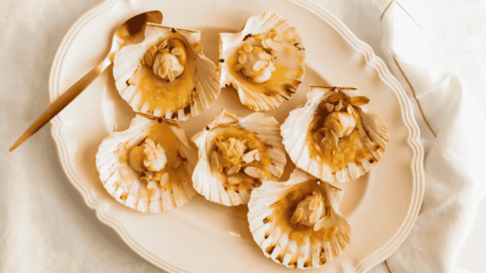 Six baked scallops topped with almonds served in their shells on a scalloped white plate with a gold spoon