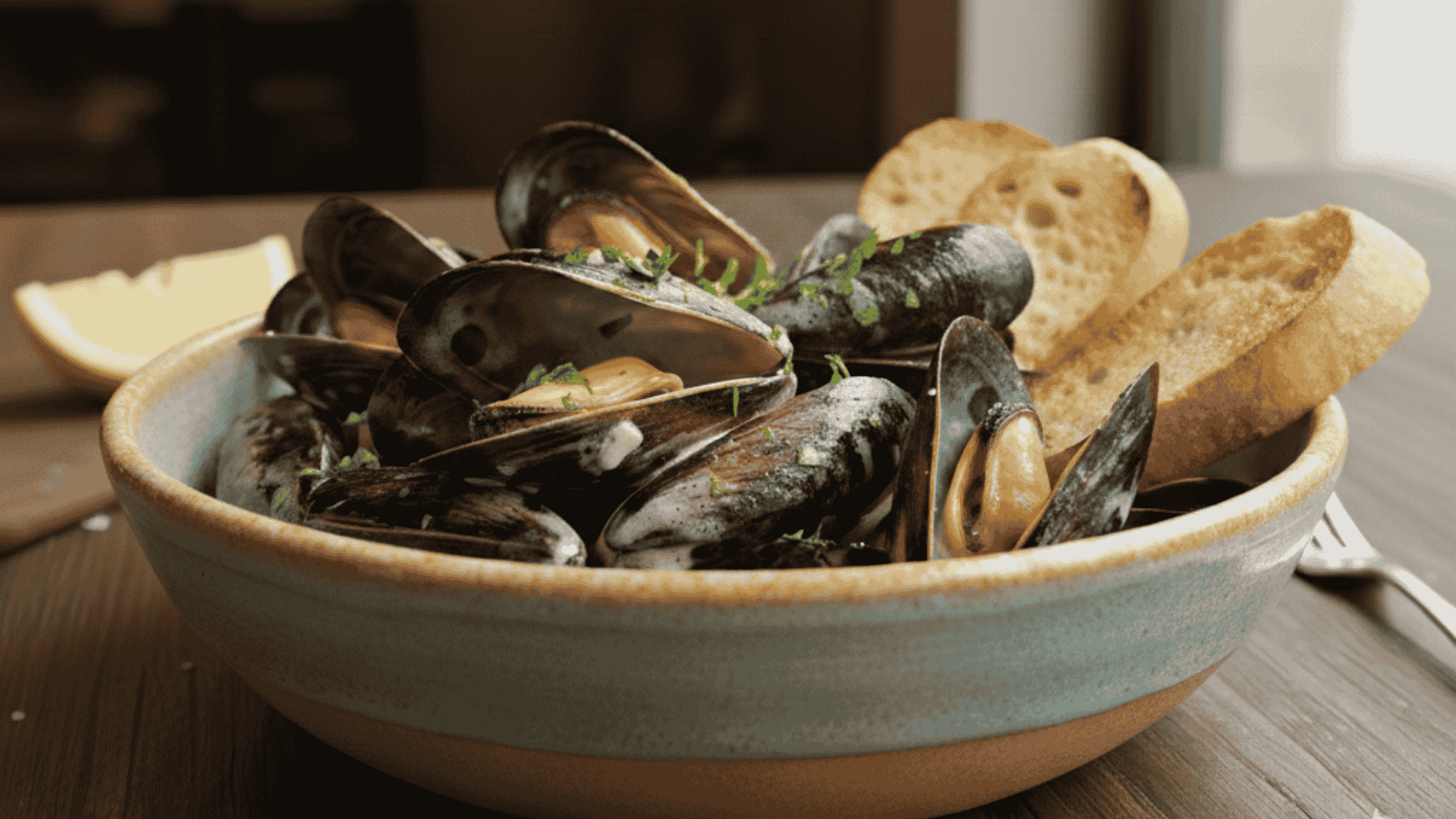 Steaming black mussels in white wine broth garnished with herbs served in a white bowl beside a slice of rustic bread