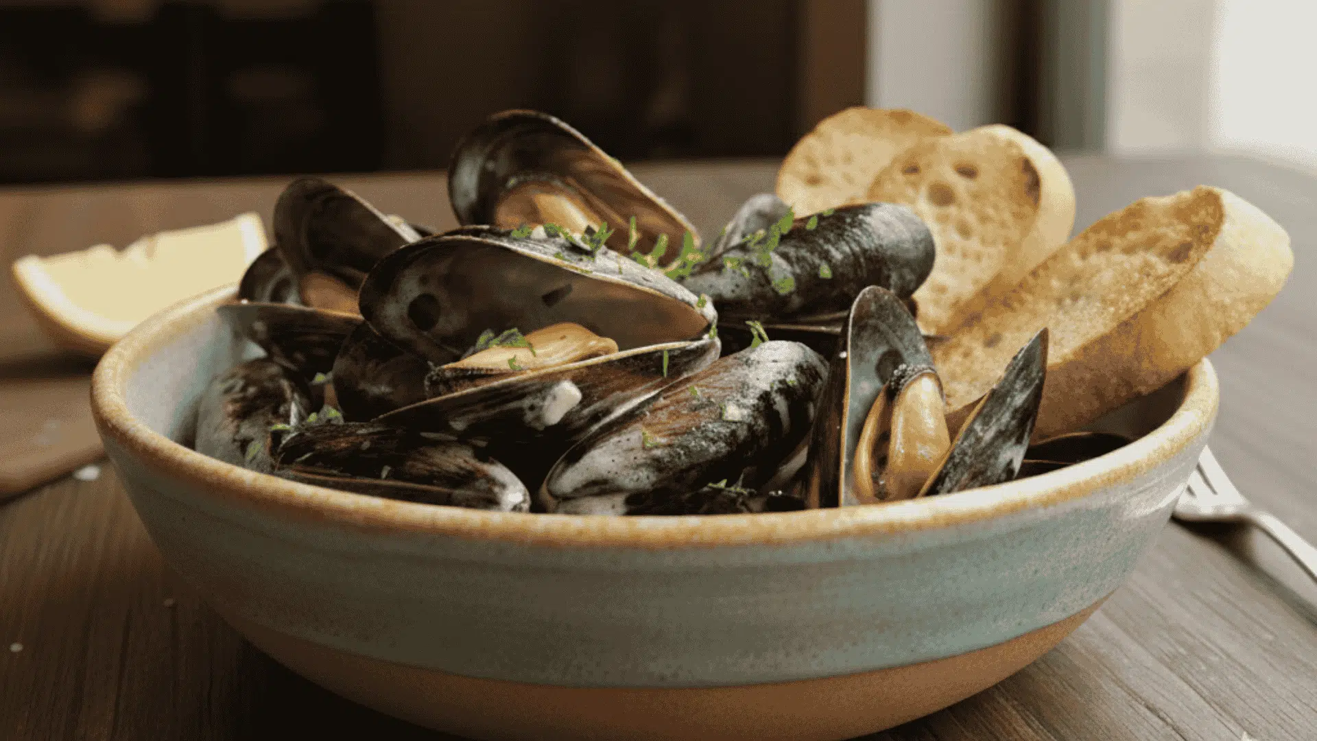 Steaming black mussels in white wine broth garnished with herbs served in a white bowl beside a slice of rustic bread