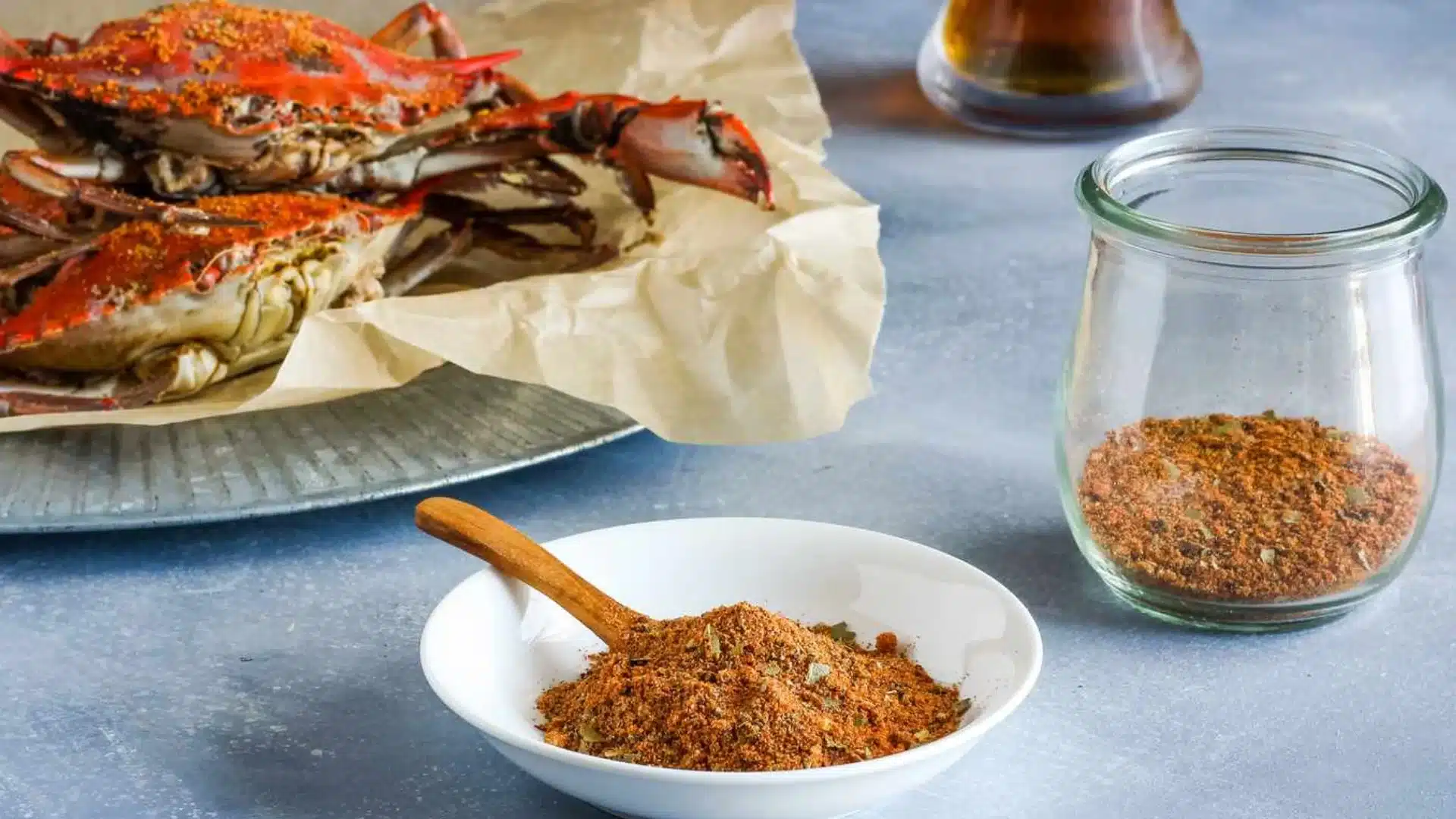 An image of steamed crabs, seasoned with a Cajun spice mix, in a bowl and jar, set on a gray surface