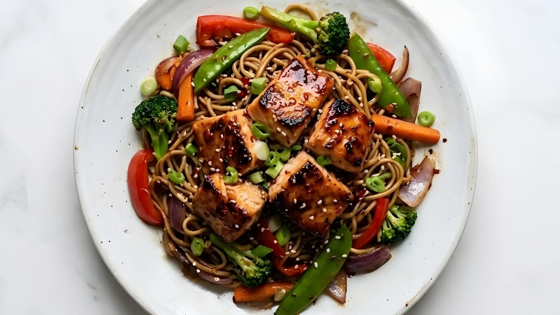 Overhead view of glazed salmon pieces and stir fry noodles with mixed vegetables on a white plate over marble surface