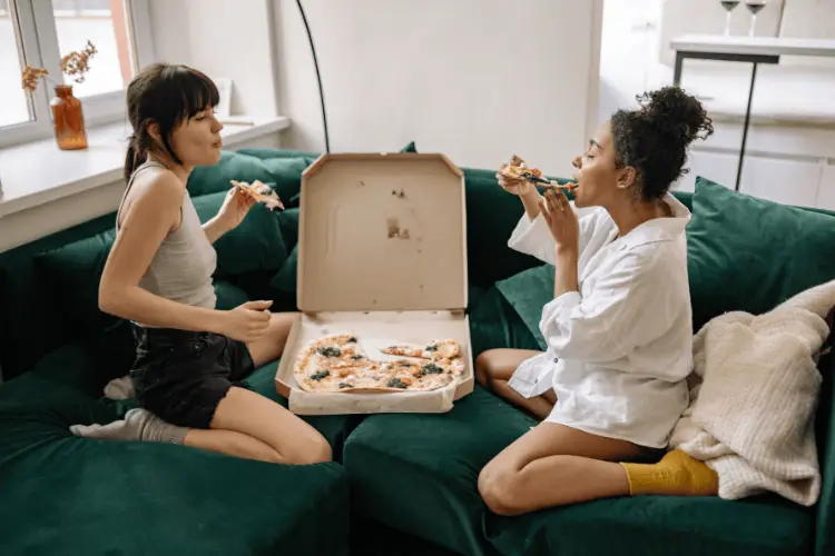 Two women enjoying pizza on a green sofa in a cozy living room setting