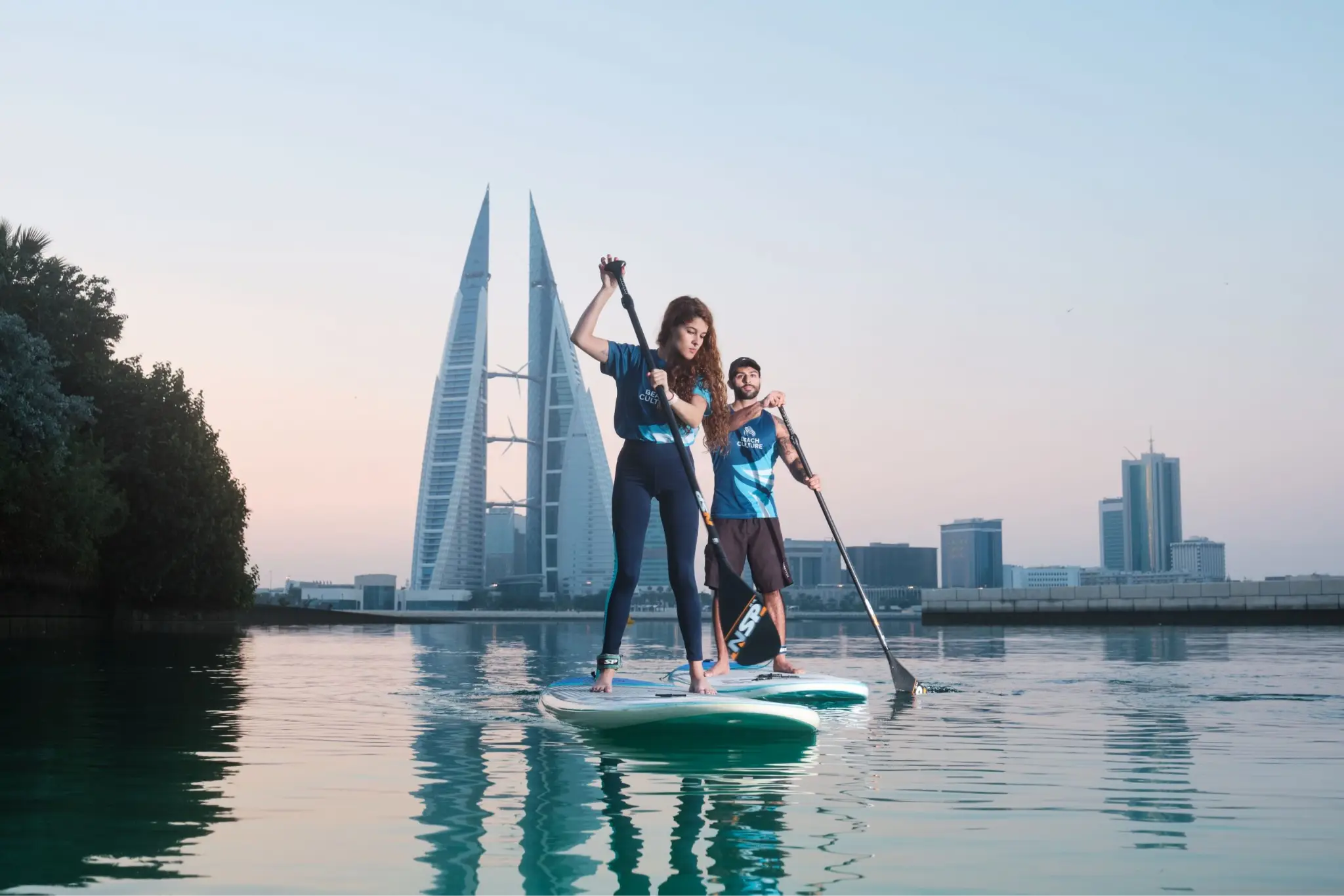 Two people paddleboarding on calm water with city skyline and modern towers in background