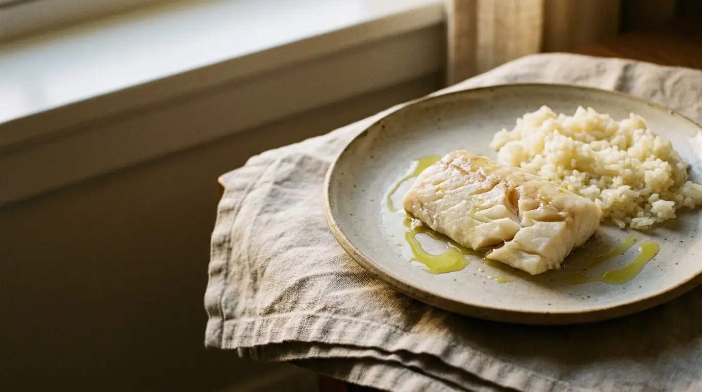 Cooked fish fillet with rice drizzled with oil on ceramic plate by window