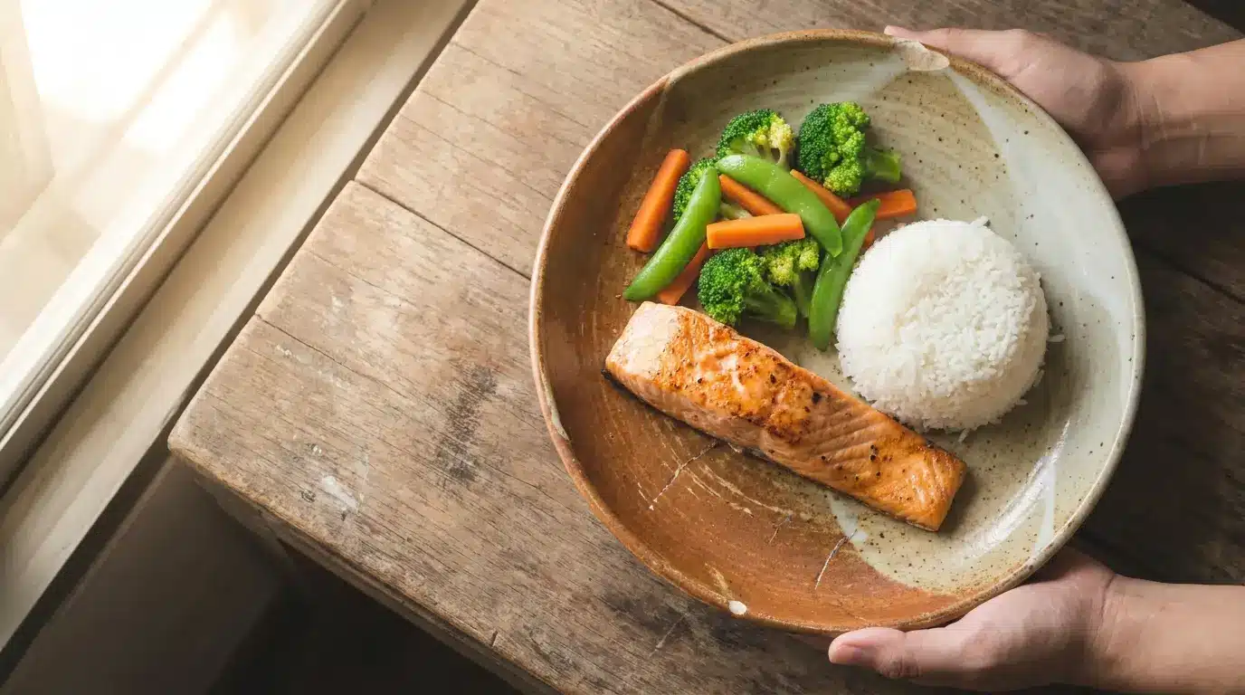 Grilled salmon with steamed vegetables and rice on rustic wooden table near window