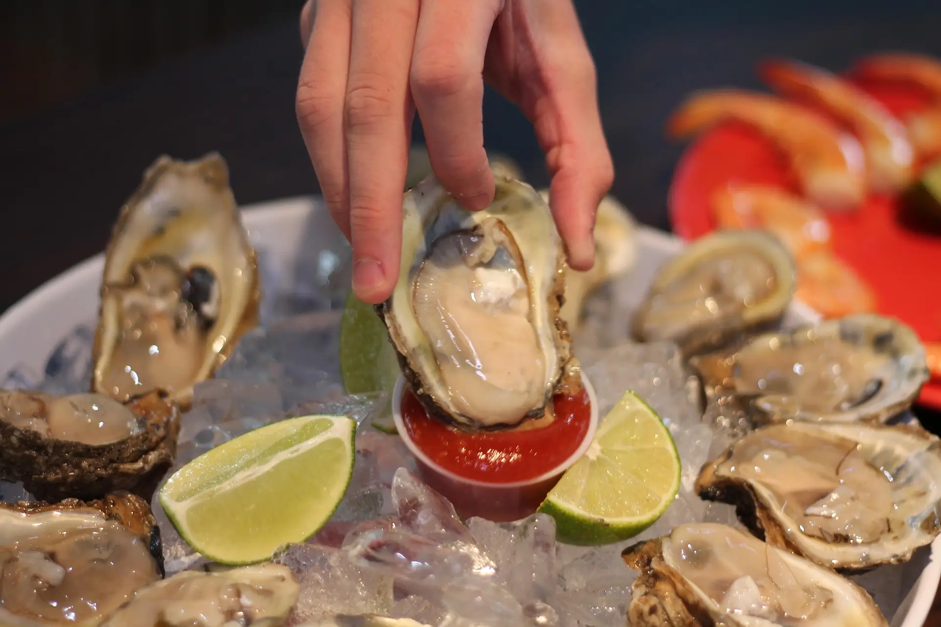 Hand holding oyster over cocktail sauce and ice with lime wedges in seafood platter
