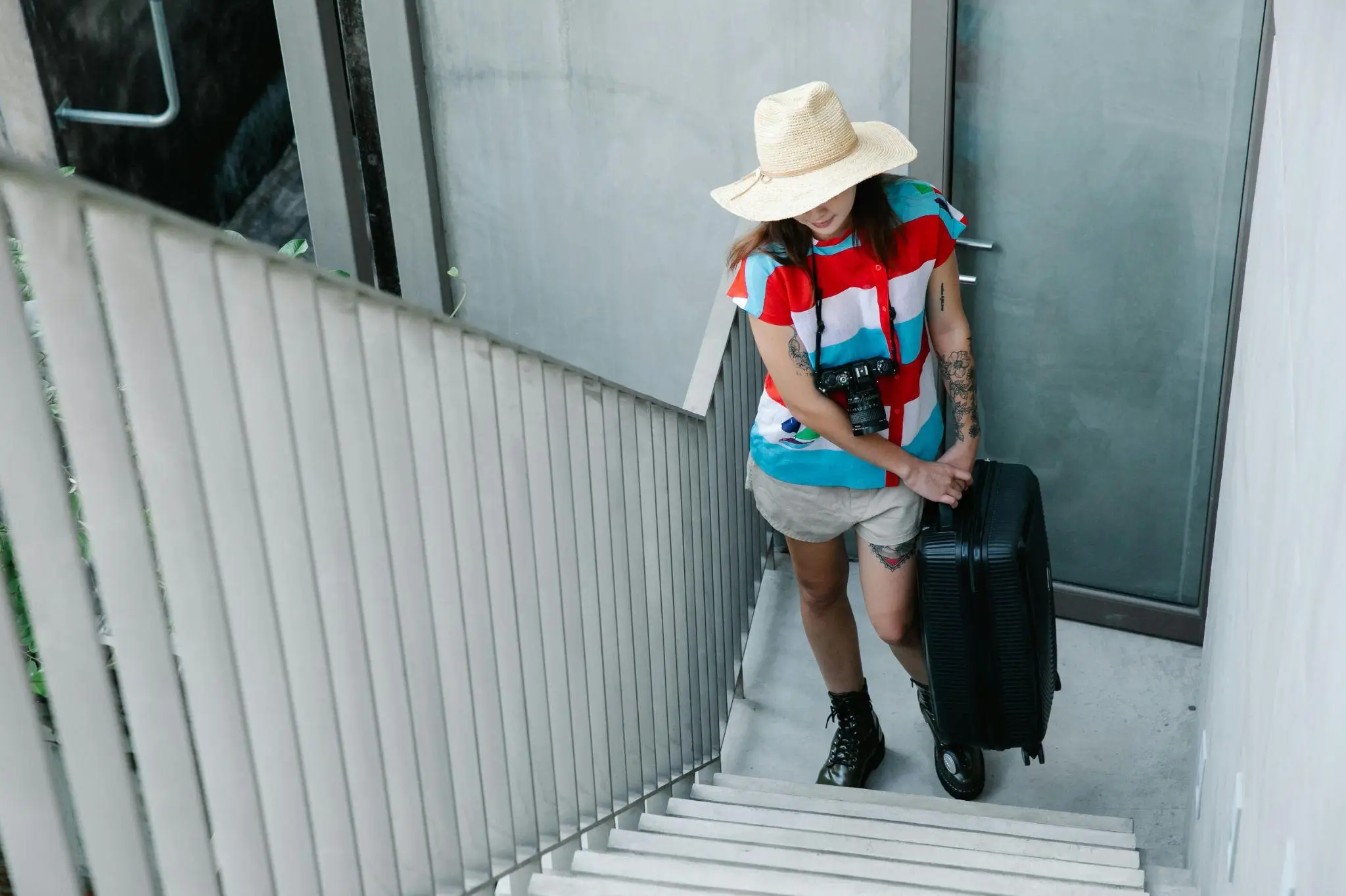 Person in bright shirt and sun hat carrying suitcase up outdoor concrete stairs