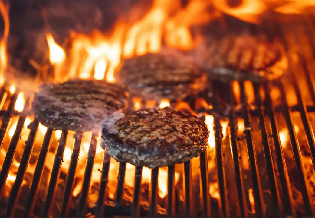 Burger patties grilling over open flames with smoke and warm lighting