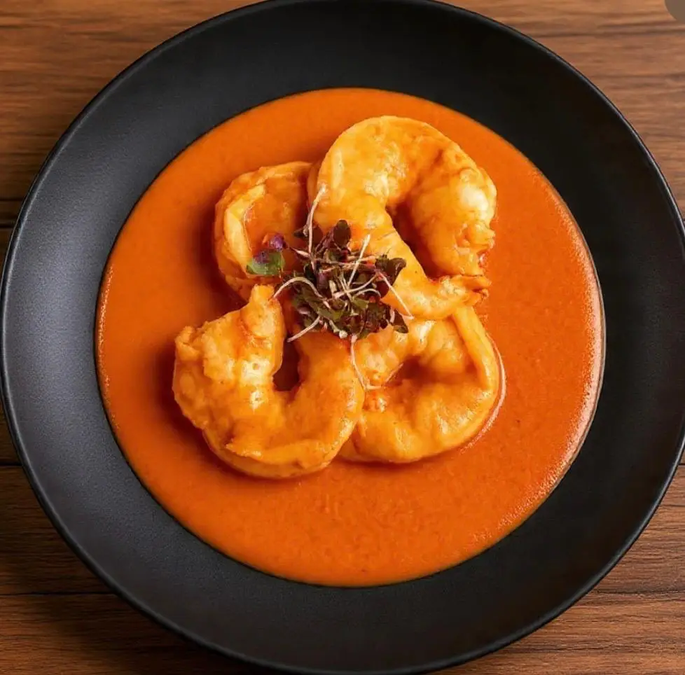Shrimp in orange sauce garnished with microgreens on black plate in warm lighting