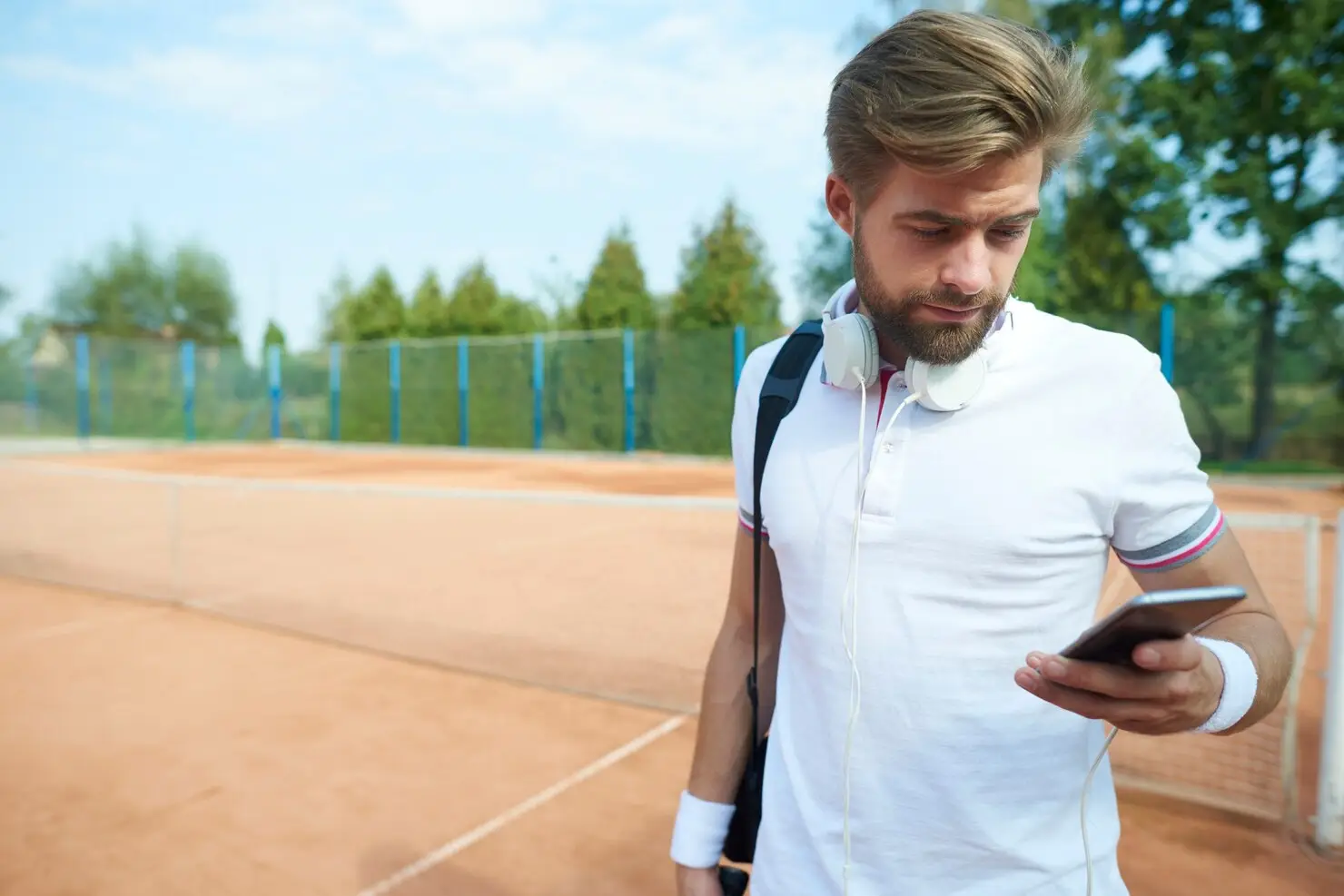 Man with headphones and backpack checking phone on outdoor tennis court in sunlight