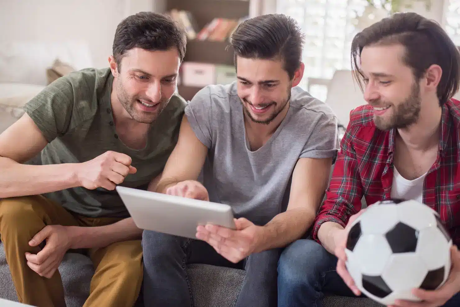 Three men sitting on a couch looking at a tablet, one holding a soccer ball