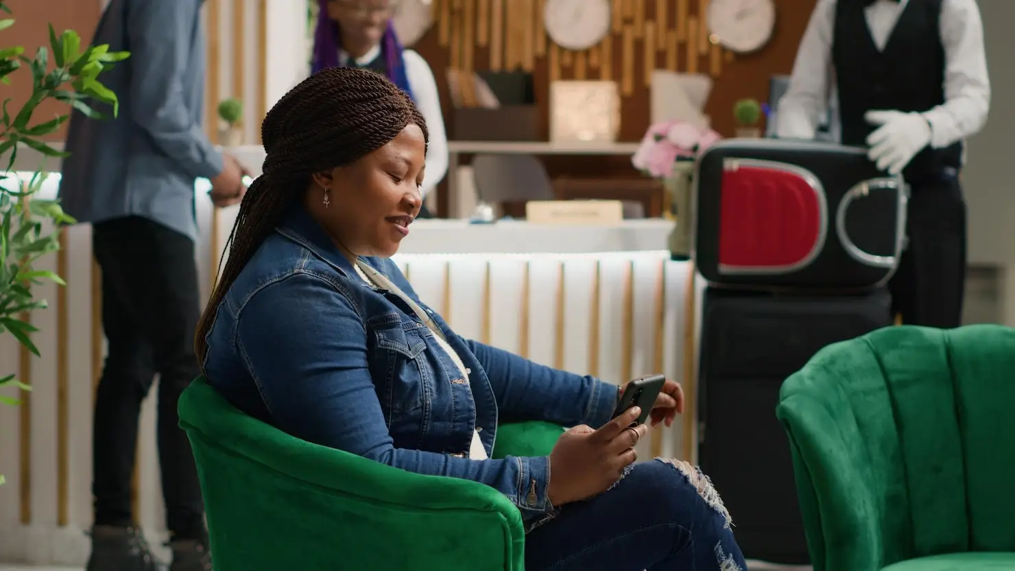 Woman in denim jacket using smartphone in modern hotel lobby with green velvet chair