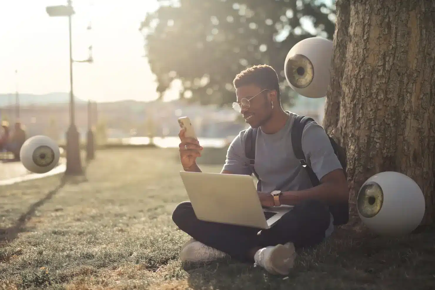 Person sitting under tree using laptop and phone, surrounded by floating eyeballs in a sunny park