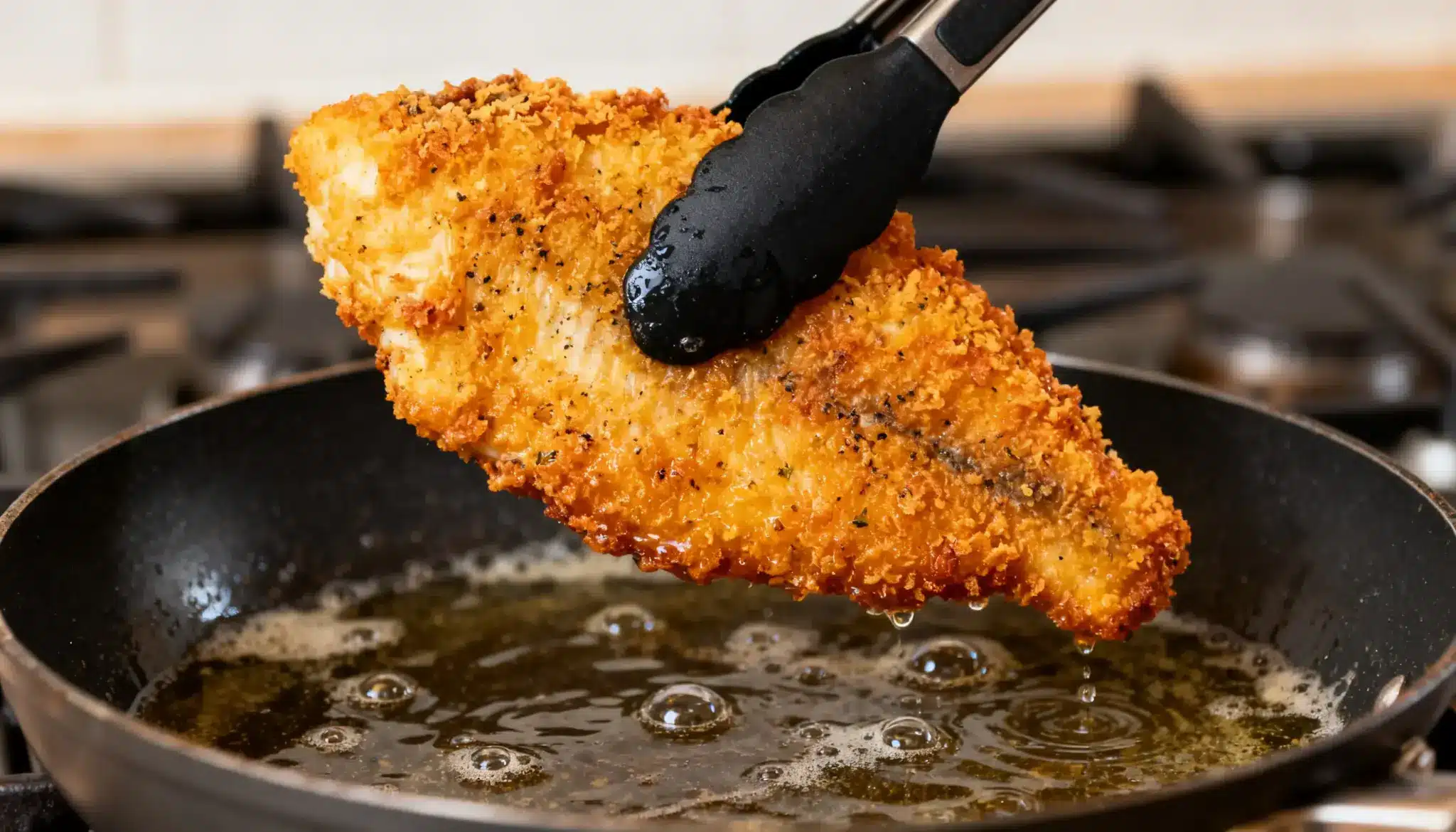 A piece of crispy breaded fish is lifted from hot oil in a frying pan with black tongs.