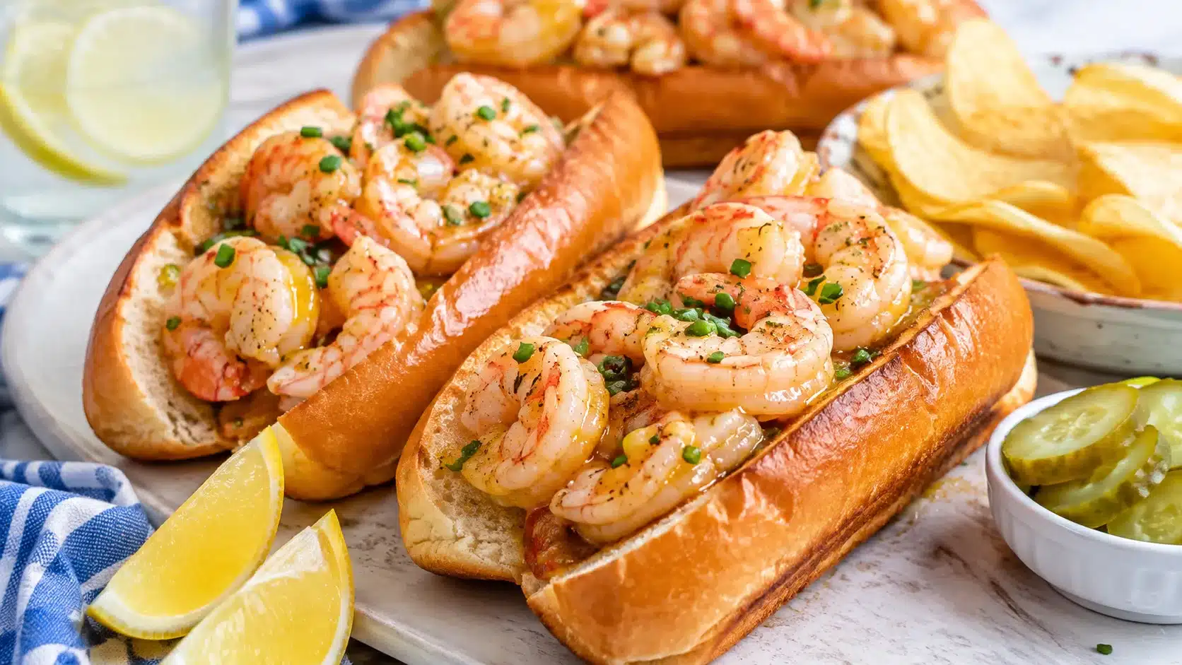 warm butter shrimp rolls in toasted buns with lemon, chives, pickles, and potato chips on a serving board