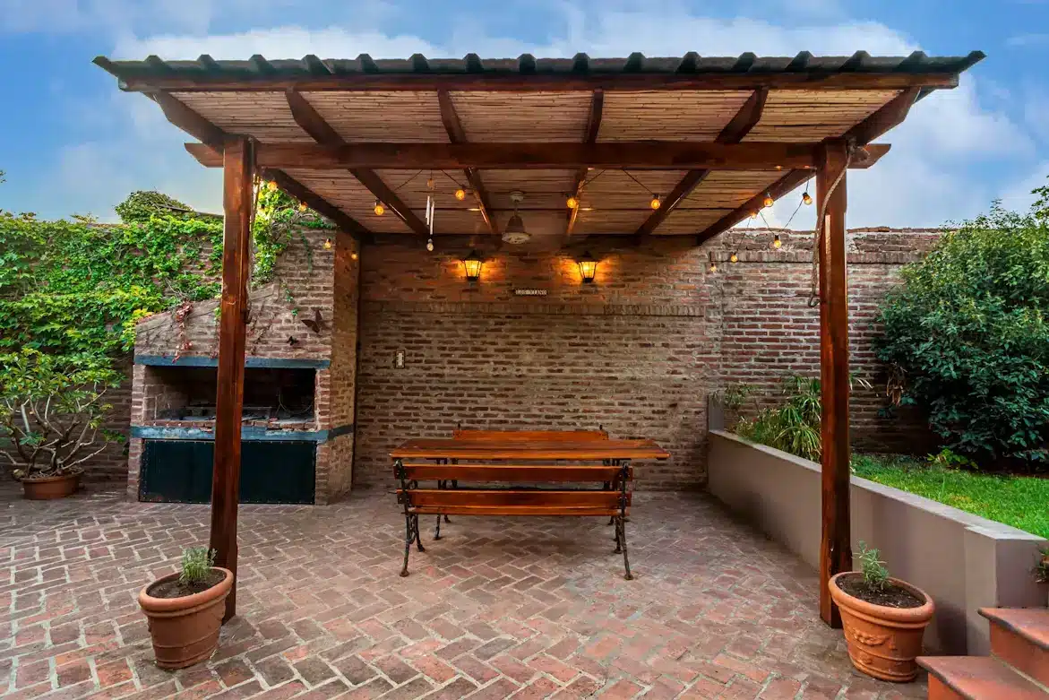 Outdoor patio with pergola, brick grill, wooden benches, and warm string lights on cobblestone floor