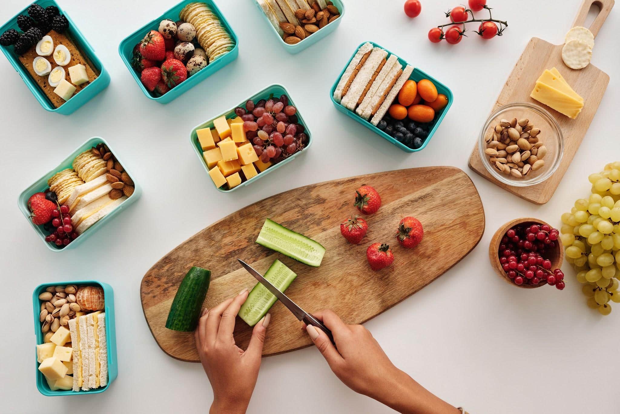 Hands slicing cucumber on wooden board with assorted snacks and fruit in teal containers