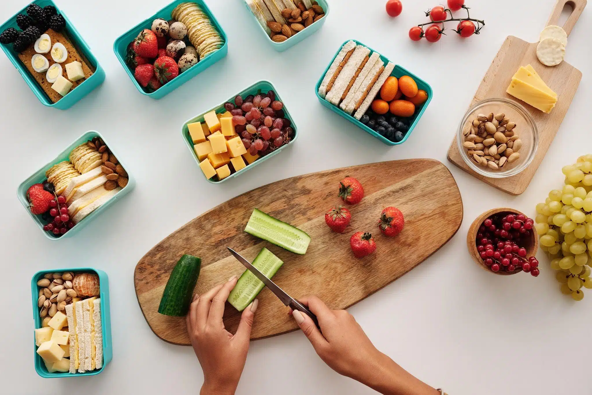Hands slicing cucumber on wooden board with assorted snacks and fruit in teal containers
